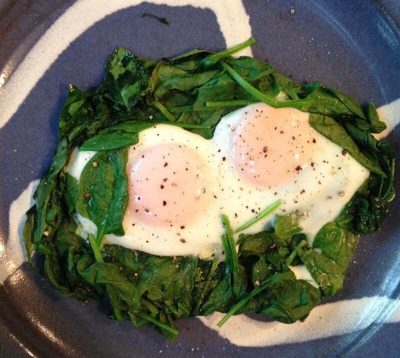 Two eggs poached and sprinkled with pepper sitting on top of a bed of wilted spinach. These are sitting in a beautiful denim blue plate with white swirls.with 