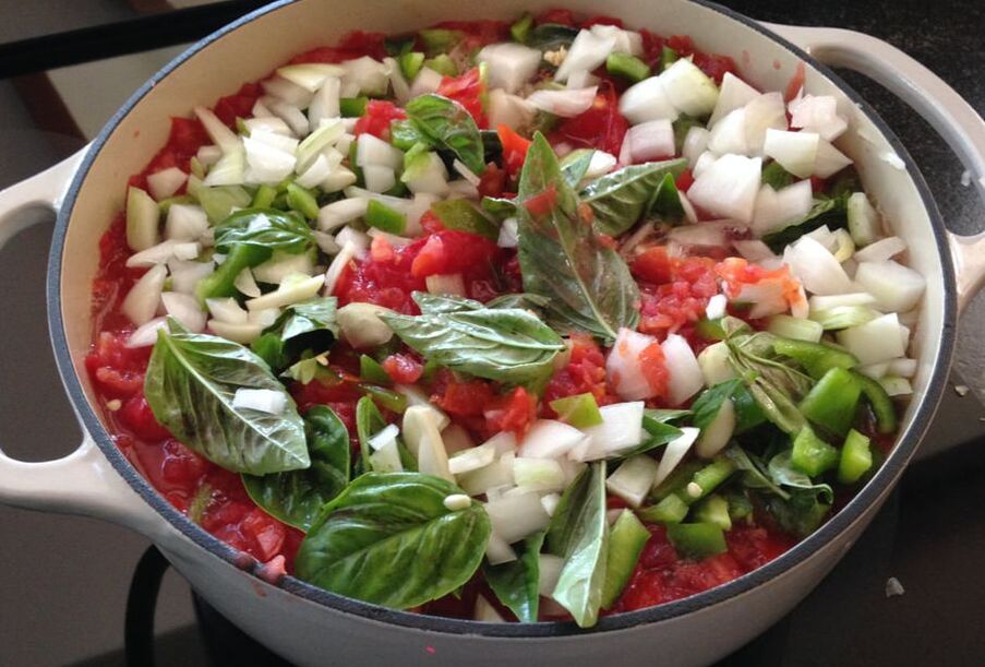 A white enamaled cast iron pot filled with a colorful combination of tomatoes, onions and topped with basil leaves.