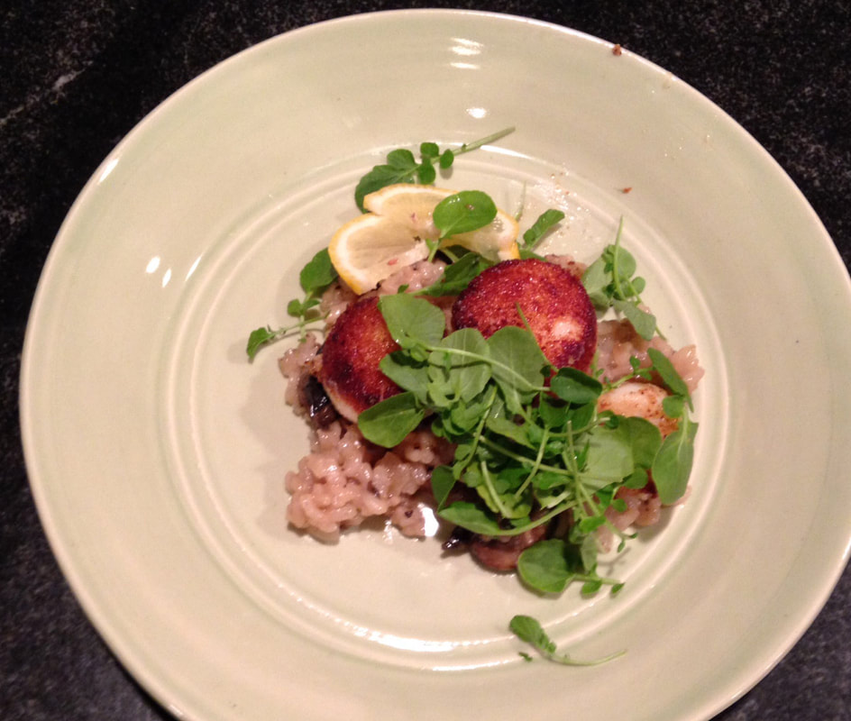 A beautiful handmade porcelain plate holding a serving of risotto and topped with seared scallops and rocket or argula; accompanied by a lemon wedge on the side.